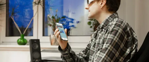 Wie der Berufsalltag blind gelingt: Eine blinde Person sitzt am Arbeitsplatz und hält ein Handy in der Hand um sich die Inhalte von einer Software vorlesen zu lassen