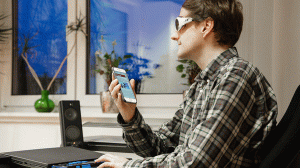 Wie der Berufsalltag blind gelingt: Eine blinde Person sitzt am Arbeitsplatz und hält ein Handy in der Hand um sich die Inhalte von einer Software vorlesen zu lassen